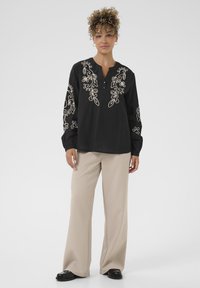 Sort bluse med detaljeret hvid blomsterbroderi på ærmerne og brystet, med henley-udskæring og lange ærmer. Sæt sammen med beige bukser.