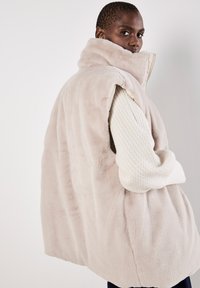 Neplederen gilet in zacht beige met een hoge col, mouwloos ontwerp en een geribde witte trui eronder. Soepele textuur en relaxte pasvorm.