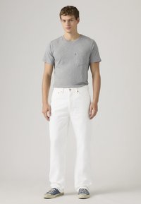 Νεαρός άνδρας που φοράει γκρι κοντομάνικη μπλούζα, λευκό τζιν παντελόνι και μπλε αθλητικά παπούτσια, στέκεται μπροστά σε ένα απλό ανοιχτό φόντο.