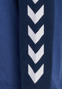 Mørkeblå sweatshirtstof med en struktureret overflade. Har hvide chevron-formede mønstre langs ærmet i en vertikal opstilling.