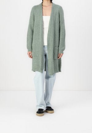 Kvinde iført lys grøn åben-front lang cardigan med lommer, hvid top, lyseblå jeans og sorte sneakers med beige såler.