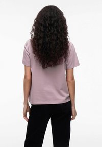 Lilla bomulds T-shirt med afslappet pasform, korte ærmer og lige kant, parret med sorte bukser. Tekstureret, naturligt krøllet hår synligt.