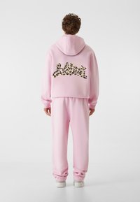 Roza dukserica in usklajene hlače z leopardjim vzorcem na hrbtni strani. Mehak material, sproščen kroj in kapuco.