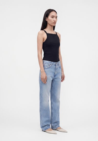 Γυναίκα που στέκεται, φοράει μαύρο αμάνικο τοπ με τετράγωνη λαιμόκοψη, ανοιχτό μπλε τζιν παντελόνι με ευθεία γραμμή και μπεζ μυτερά μπαλαρίνια, σε λευκό φόντο.