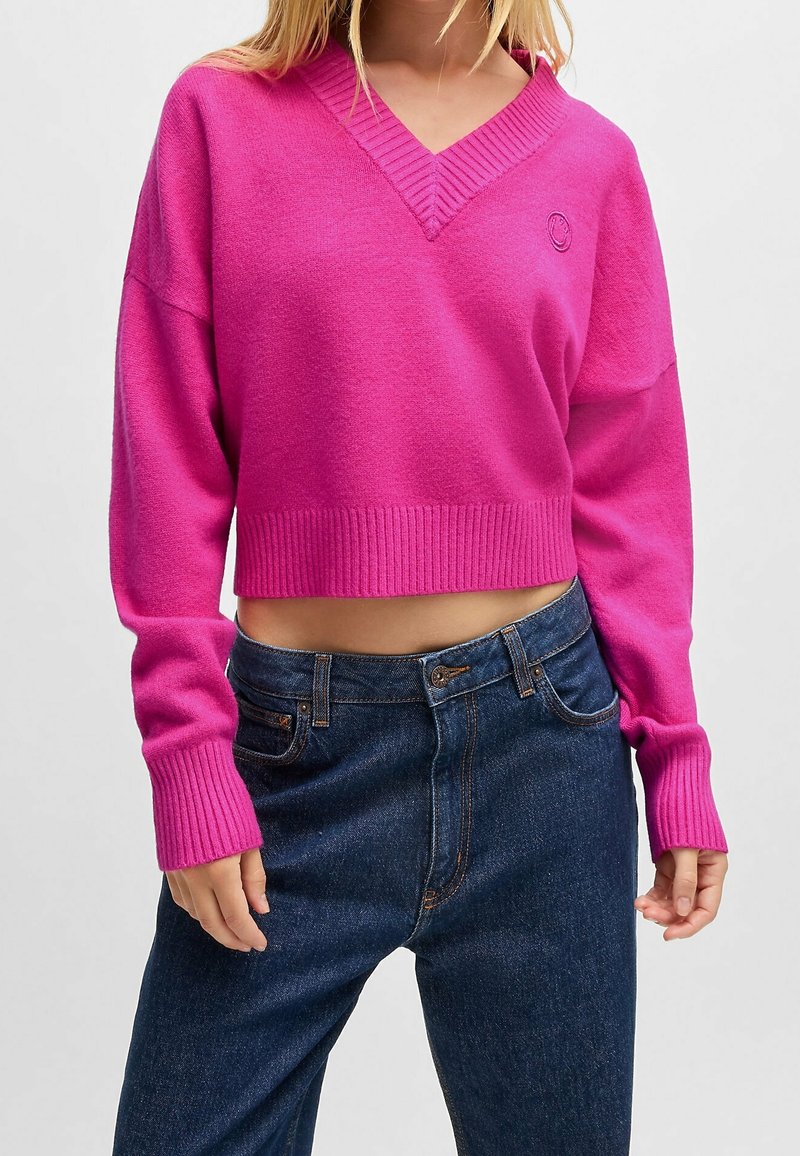 Femme portant un pull court rose vif à col en V avec des manches longues et un jean taille haute bleu foncé sur un fond uni.