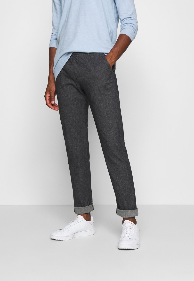 Mężczyzna w ciemnoszarych podwiniętych spodniach, jasnoniebieskiej koszuli z długim rękawem i białych trampkach, stoi z jedną ręką w kieszeni.