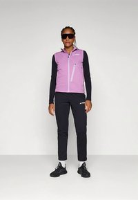 Violet zip-up liemenė ant juodos ilgomis rankovėmis marškinėlių, derinama su juodomis kelnėmis ir juodais sportbačiais. Ypatybės apima užtrauktuką kišenėje ant liemenės.