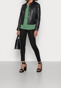 Svart läderjacka, grön topp, svarta mockaleggings och svarta klacksandaler med blockklack. Håller en liten svart handväska.