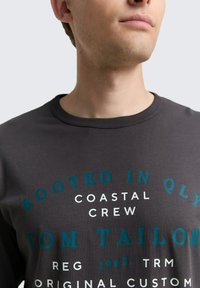 Dunkelgraues Baumwoll-Crewneck-T-Shirt mit auffälligem weißen und türkisfarbenem Text, der Markendetails und ein rundes Design zeigt. Weiche Textur, lässige Passform.