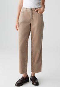 Pantaloni beige a gamba dritta con una superficie liscia, quattro tasche e chiusura a pulsante, abbinati a mocassini scuri.