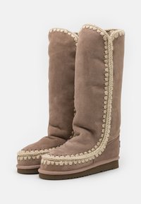 Bottes en daim marron hauteur genou avec des coutures décoratives beiges le long des coutures et une semelle texturée pour une bonne adhérence.