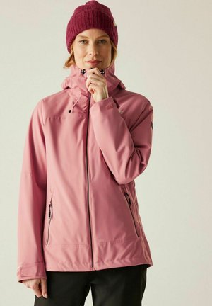 Kobieta w bordowej dzianinowej czapce i różowej kurtce z kapturem na suwak, stojąca na tle jasnego, neutralnego tła.