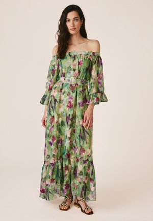 Donna che indossa un vestito lungo verde floreale con spalle scoperte, una cintura e sandali con decorazioni, in piedi davanti a uno sfondo semplice.