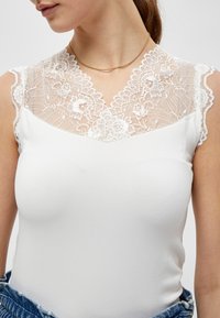 Top bianco senza maniche con corpetto in pizzo caratterizzato da motivi floreali, un bordo ondulato e una finitura in tessuto liscia. Una collana dorata completa il look.