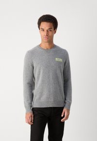 Pilkas megztinis su apykakle, pagamintas iš švelnios vilnos, su žaliu logotipo užtrauktuku ant krūtinės ir gipiūro rankovėmis bei dugnu. Derinamas su juodomis kelnėmis.