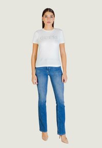 T-shirt bianca aderente con scritta "GUESS" in argento, abbinata a jeans denim blu. Il modello indossa scarpe con tacco alto beige chiaro. Design semplice.