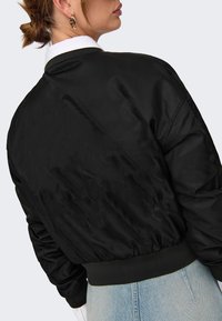 Black bomber jacket with a smooth, shiny texture, ribbed cuffs, and waist. Features a classic design, worn over a white collared shirt.