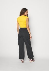 Tight passform i gul bomullstopp som matchas med lösa svarta byxor med fickor, samt maroon och vita sneakers. Bakifrån mot en vit bakgrund.