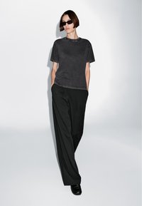 T-shirt grigia a maniche corte con scollatura rotonda, abbinata a pantaloni larghi neri. Il modello indossa occhiali da sole scuri e scarpe nere.