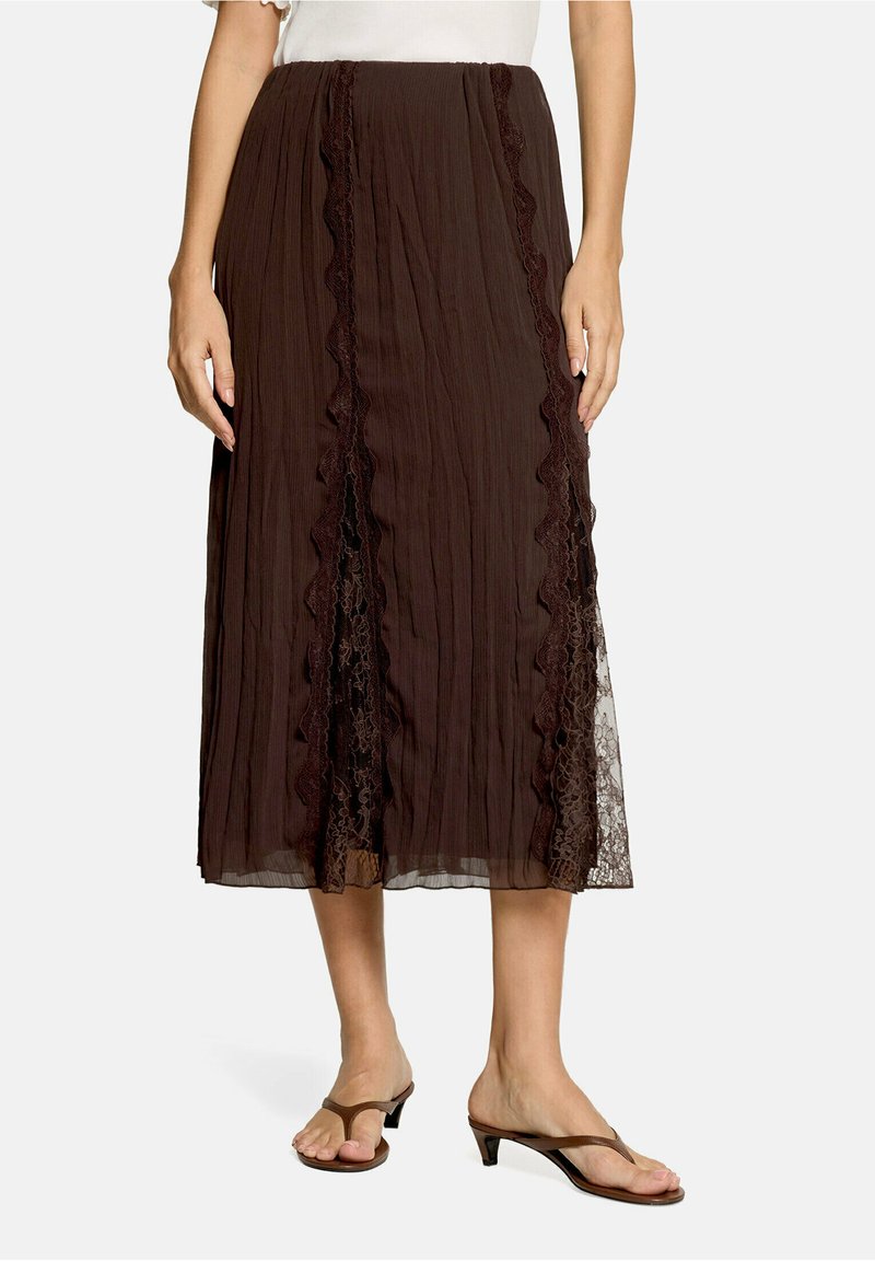 Femme portant une jupe midi marron avec des panneaux en dentelle verticale et des détails à volants, assortie à des sandales à talons marron.