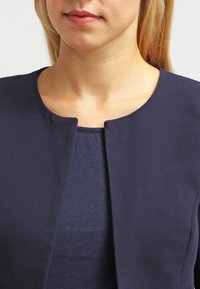 Veste bleu marine avec un col rond et sans fermetures, réalisée en tissu lisse ; associée à une chemise assortie bleu marine en dessous.