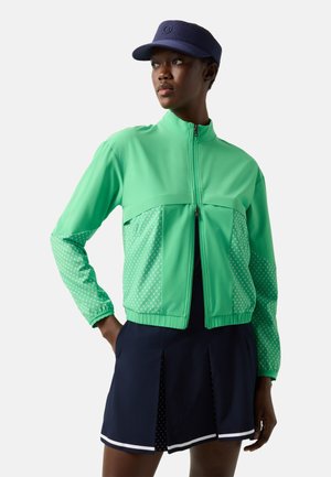 Žena v zelené bundě na zip s vzorovanými rukávy, tmavě modré sukni s bílým lemem a tmavě modrým kšiltu čeká, zatímco se dívá stranou.