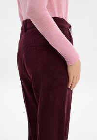 Bordeaux corduroy broek met smalle verticale strepen, voorzien van een fitted taille en zijzakken. Lichtroze lange mouw top gedragen.