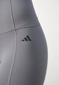 Leggings sportivi grigi realizzati in un tessuto morbido e elastico, con una sezione superiore testurizzata. Presentano un logo nero a tre righe sul lato.