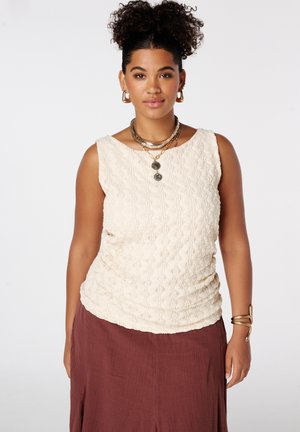 Vrouw met krullend haar draagt een crèmekleurige, mouwloze top met textuur, een bordeaux rok, gouden oorringen, gelaagde kettingen en een armband, kijkt recht vooruit.