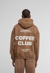 Sudadera de algodón marrón con un gran texto blanco en la espalda que dice "COFFEE CLUB" y un texto más pequeño arriba y abajo. Cuenta con capucha y un ajuste relajado.