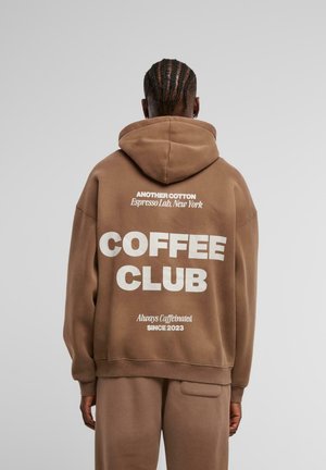 Felpa in cotone marrone con grande testo bianco sul retro che recita "COFFEE CLUB" e testo più piccolo sopra e sotto. Presenta un cappuccio e una vestibilità rilassata.