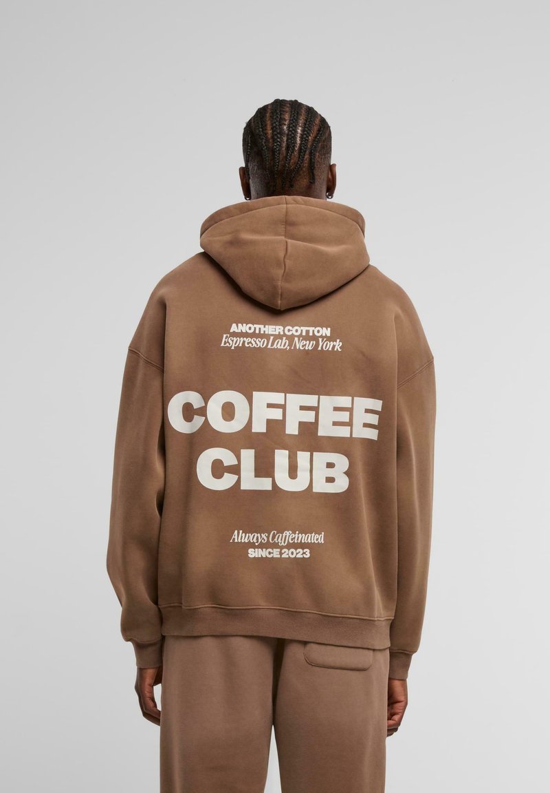 Sudadera de algodón marrón con un gran texto blanco en la espalda que dice "COFFEE CLUB" y un texto más pequeño arriba y abajo. Cuenta con capucha y un ajuste relajado.