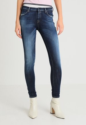Vaqueros ajustados de color azul oscuro con un lavado desgastado, cinco bolsillos y trabillas para cinturón, combinados con botas blancas hasta el tobillo y una blusa rosa claro.