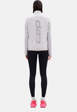 Hellgraue Sportjacke mit hohem Kragen und großem Motiv auf dem Rücken. Schwarze Leggings mit Punktemuster und pinke Schuhe.