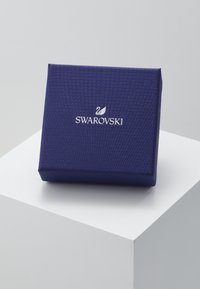 Swarovski koruloot sügavale sinisele tekstuuritud materjalile, millel on reljeefne muster ja metallik hõbe Logo ees. Ruudukujuline.