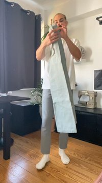 Šviesiai mėlynos medžiagos kelnės laikomos rankoje, turi tiesią kirpimo dizainą. Nešiojamos pilkos kelnės ir baltos kojinės, matosi medinės grindys.