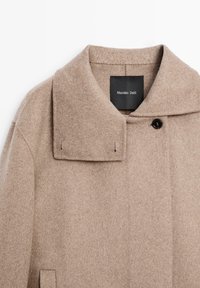 Beige ovčí kabát s velkým límcem, dvouřadým zapínáním a jemnou strukturou. Label viditelný uvnitř. Snadný a funkční design.