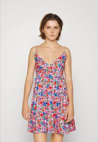 Mulher vestindo um vestido sem mangas, com decote em V, com padrões florais em rosa, vermelho, azul e verde sobre um fundo branco.