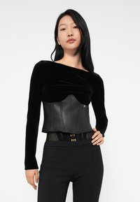 Black velvet long-sleeve top with draped neckline and leather corset overlay. Accented with gold hardware and fitted design, paired with black pants.