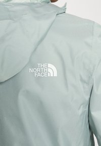 Veste bleu clair, résistante à l'eau, avec capuche. Présente le logo "The North Face" en blanc au dos. Texture lisse et design fonctionnel.