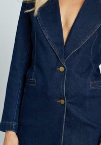 Blazer in denim di colore blu scuro con rever a punta e due tasche frontali. Presenta bottoni oro e cuciture a contrasto per un tocco di dettaglio in più.