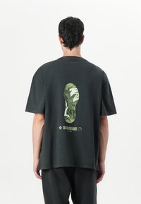 T-shirt en coton noir avec un grand graphique de semelle de chaussure verte et blanche au dos, avec des détails textuels en bas. Manches courtes, coupe décontractée.