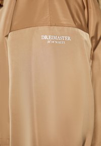 Beige jacka tillverkad av ett slätt, lätt tyg. Har vit logotext och koordinater. Designen är enkel med en matt yta.