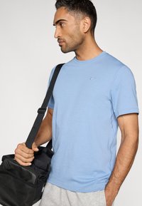 Ανοιχτό γαλάζιο κοντομάνικο μπλουζάκι με μικρό λογότυπο Nike; λεία υφή; φοράει με μαύρη τσάντα χιαστί; ορατά γκρι σορτς.