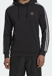 Svart hoodie av mjuk tyg, med vita tre-stripes detaljer på ärmarna, framfickan och en liten orange Adidas-logotyp på bröstet.
