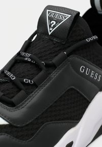 Black sneaker with textured mesh upper, smooth synthetic leather accents, white sole, elastic laces, and prominent "GUESS" logo on side.