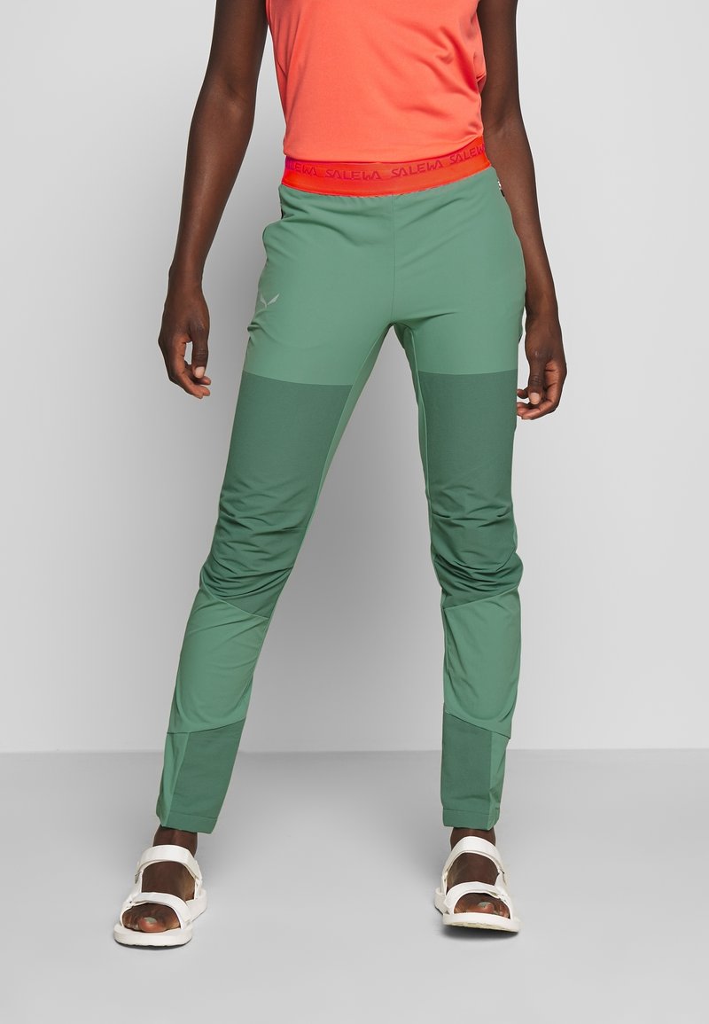 Personne portant un pantalon bicolore vert, un haut orange et des sandales blanches, debout devant un fond gris uni.