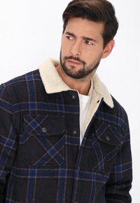 Rutete jakke i sort, blå og rød farveskema med en blød, cremefarvet pelskrave og to brystlommer med trykknaplukning.