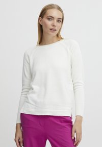 Pull en tricot blanc avec un col rond, des poignets côtelés et des fentes sur les côtés, associé à un pantalon rose. Texture lisse, coupe décontractée.