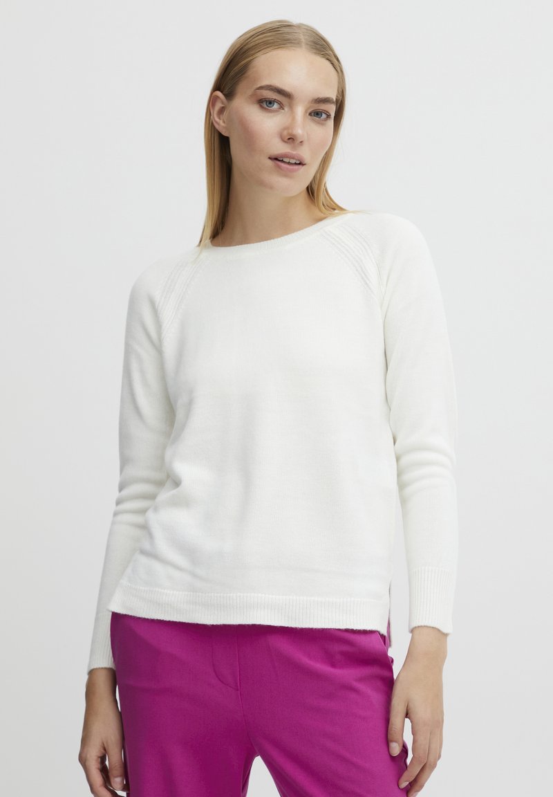 Pull en tricot blanc avec un col rond, des poignets côtelés et des fentes sur les côtés, associé à un pantalon rose. Texture lisse, coupe décontractée.